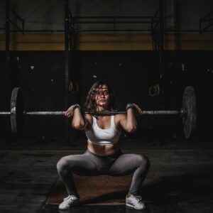 A strong female weightlifter squats with a barbell in a dimly lit gym, showcasing power and fitness.